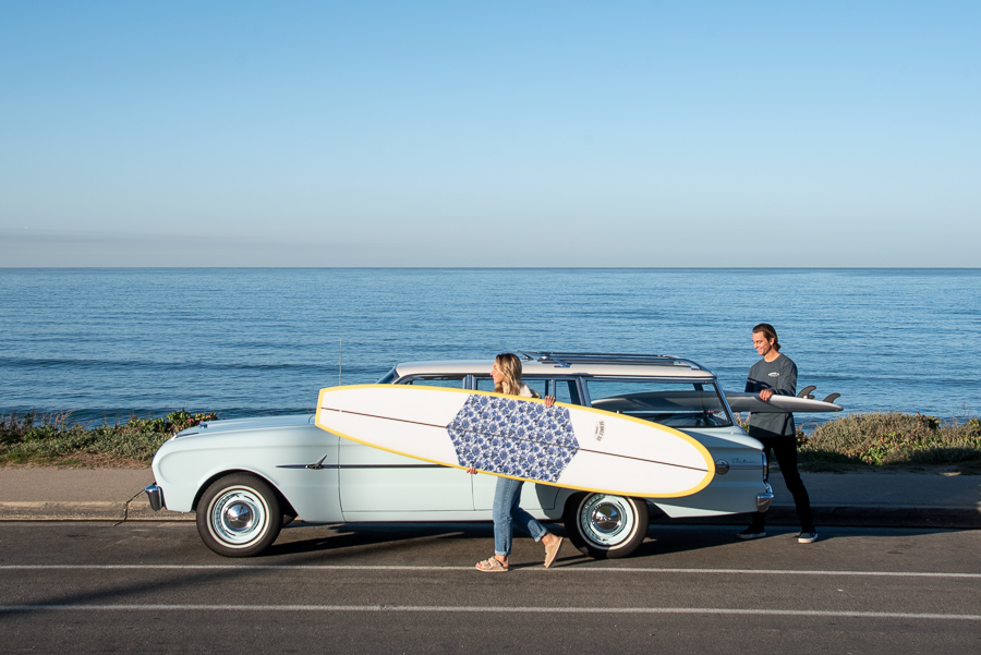 Woman carrying a longboard next to vintage car and ocean