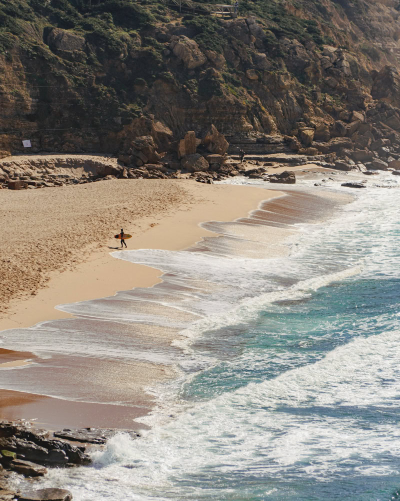 surfer walking on beach along dramatic portugal bluffs